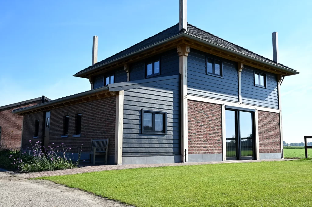 Hooiberg met donkere houten gevels, bakstenen onderbouw en lage zijvleugel naast een gazon.