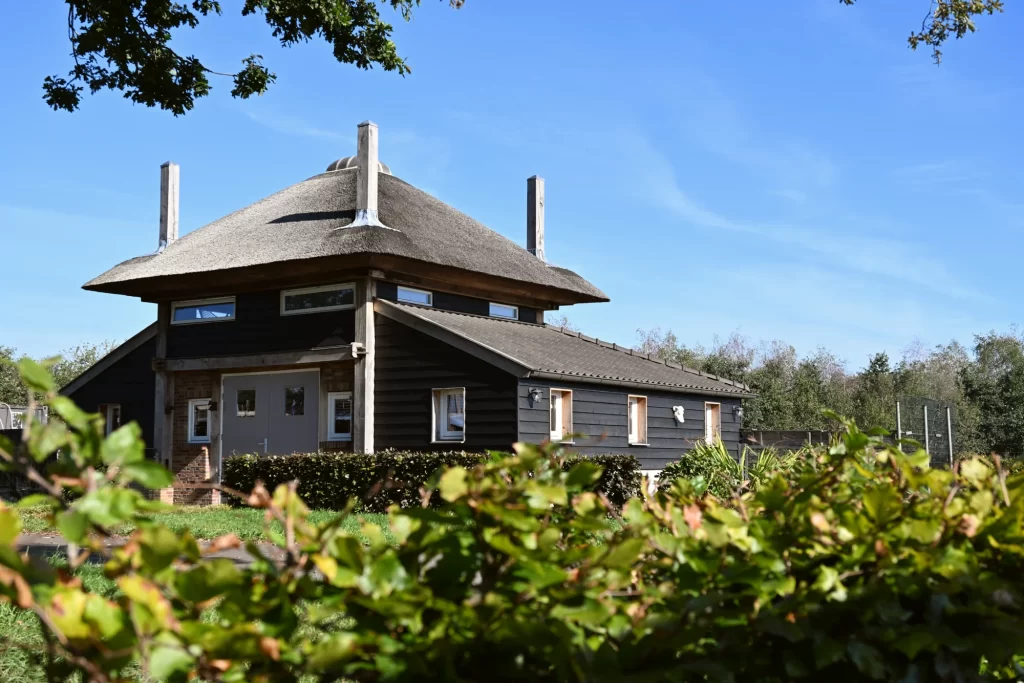 Hooiberg met rieten kap, vier uitstekende roeden en een lage aangebouwde vleugel, achter een groene haag.