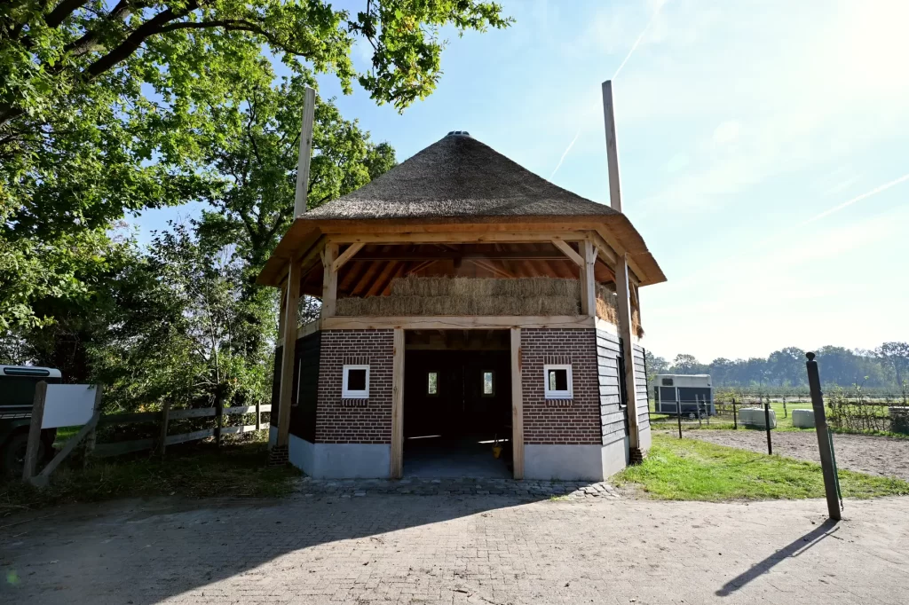 Voorzijde van een hooiberg met rieten kap, uitstekende roeden en houten deuren boven een bakstenen onderbouw.